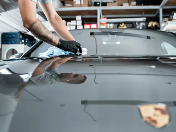 Мастер в чёрных перчатках наносит керамику микрофиброй на капот серого Porsche поверх защитной плёнки