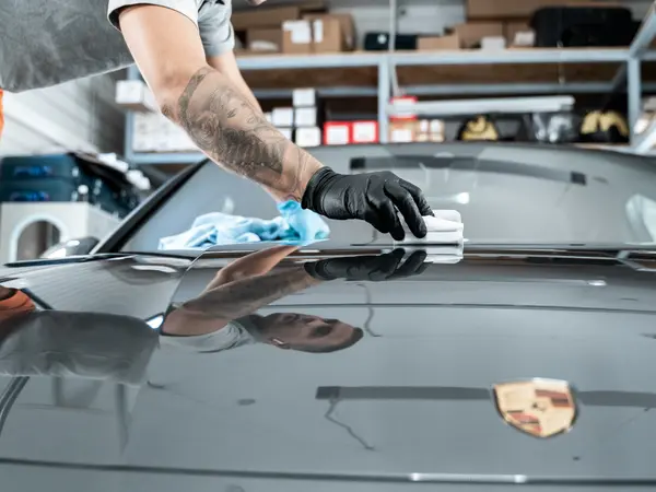 Мастер в чёрных перчатках располировывает жидкое стекло микрофиброй на капоте серого Porsche
