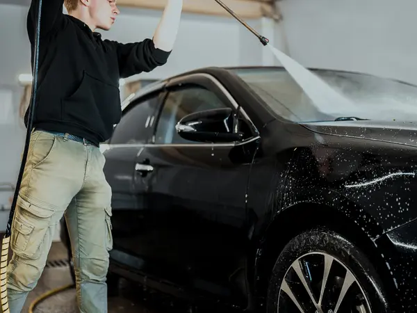 Мастер моет чёрный автомобиль аппаратом высокого давления в боксе детейлинг-центра