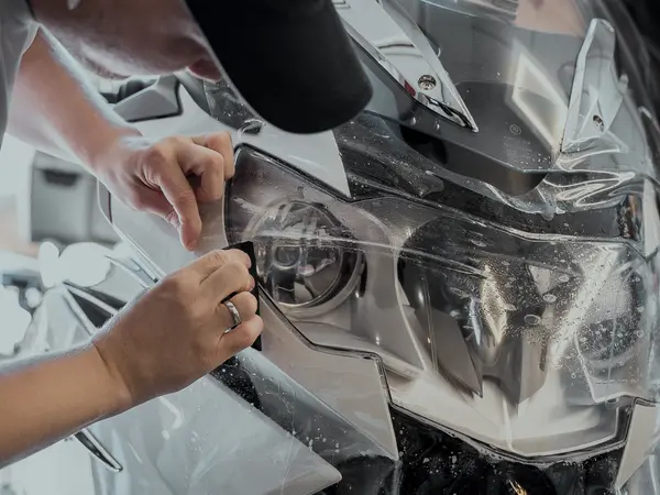 Мастер аккуратно наклеивает прозрачную защитную плёнку на фару автомобиля вручную