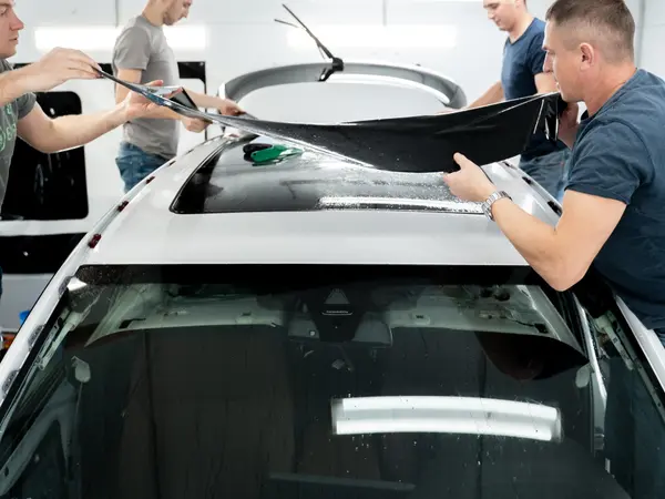 Четверо мастеров укладывают большой лист защитной плёнки на крышу белого автомобиля