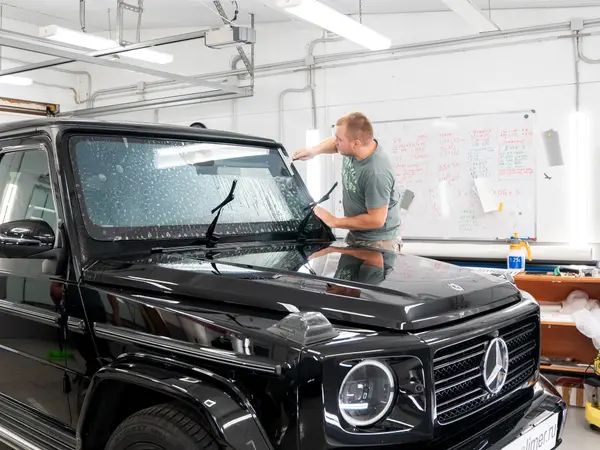 Мастер наклеивает защитную плёнку на лобовое стекло чёрного Mercedes-Benz G-Class