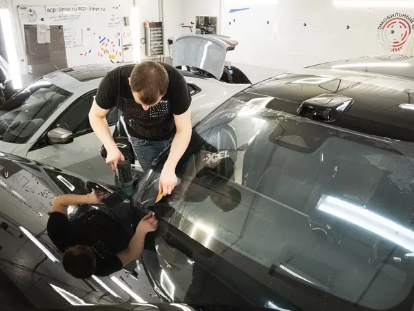 Два мастера укладывают защитную плёнку на лобовое стекло чёрного автомобиля с мыльным раствором