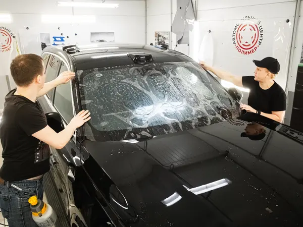 Два мастера разглаживают защитную плёнку на лобовом стекле тёмного автомобиля в цехе АЦ Полимер
