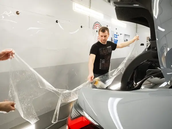 Два мастера растягивают лист защитной плёнки перед нанесением на заднюю часть автомобиля