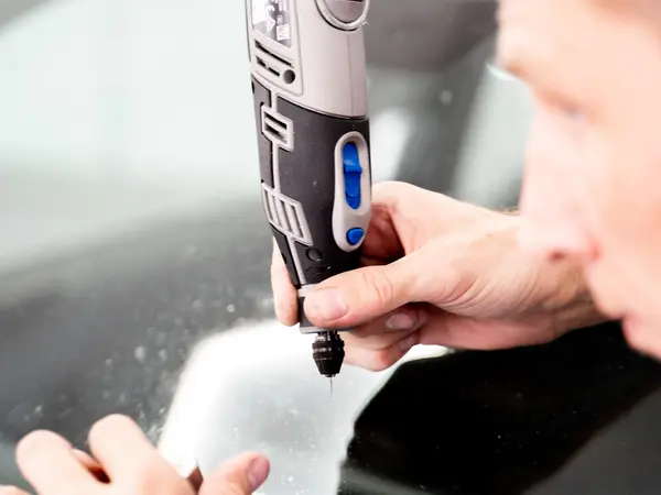 Мастер сверлит отверстие на конце трещины лобового стекла дремелем для остановки распространения