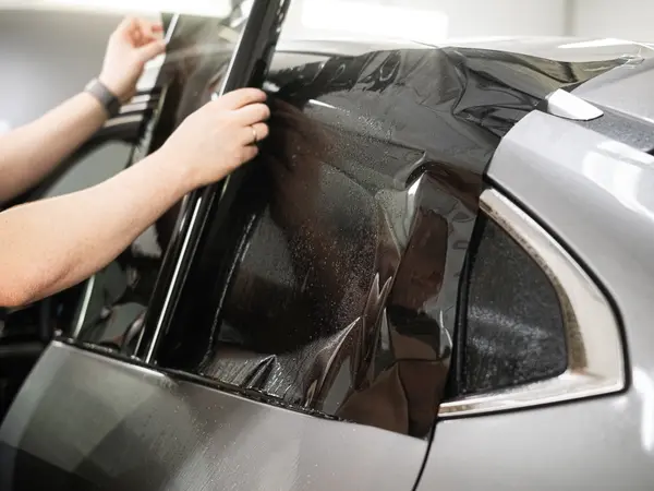 Мастер укладывает атермальную тонировочную плёнку на заднее боковое стекло серебристого автомобиля